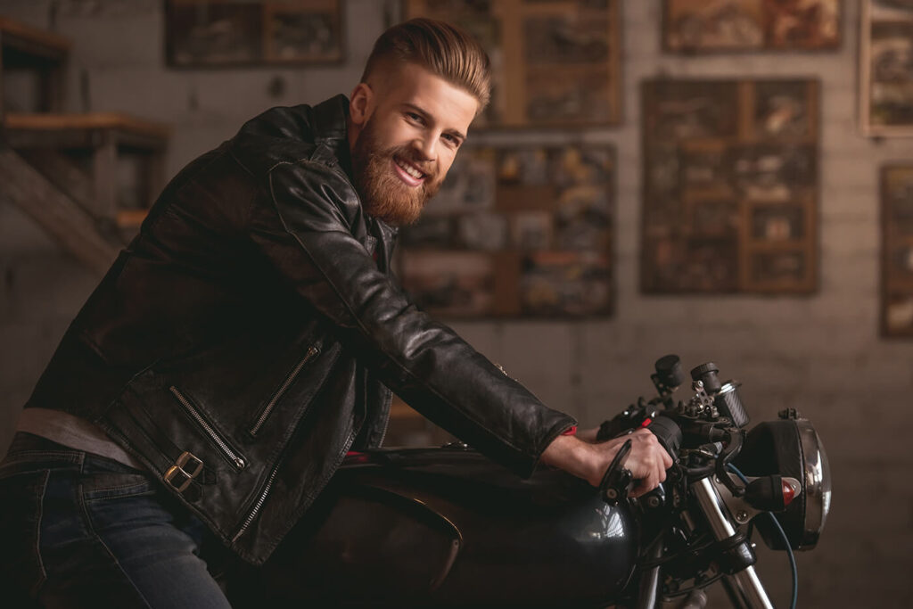 Bärtiger Mann in Lederjacke stützt sich in einer Werkstatt auf einen Café Racer mit Stummellenker